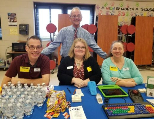 Three people sitting at a table with water bottles, information brochures and keyboards. One person is standing behind them with his arms resting on the shoulders of the people sitting in front of him. Santa Rosa County Youth Transition Fair