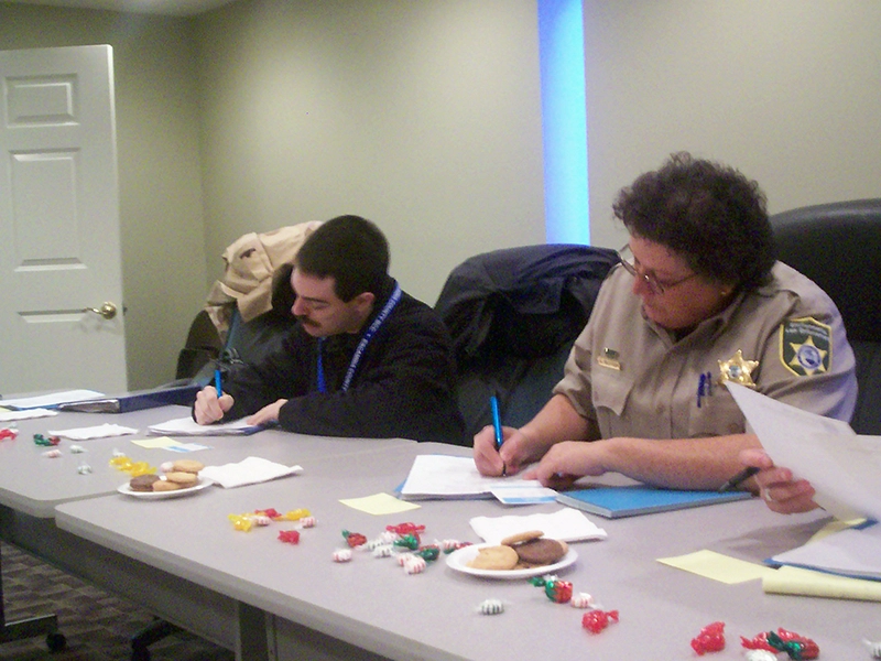 Two community members seated atparticipating in a group activity, completing paperwork while enjoying camaraderie and snacks of candies and cookies.