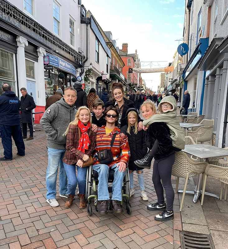 Elis in a wheelchair surrounded by family in a busy street (no cars only pedestrians)