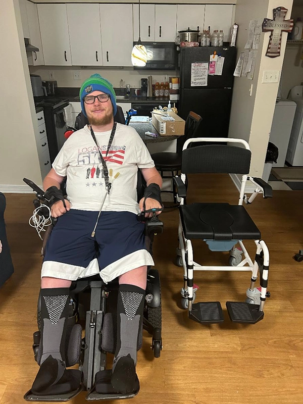 Logan seated in a powered chair next to the durable medical equipment, a Roll in Shower chair, he utilizes for activities of daily living that he received from CIL.