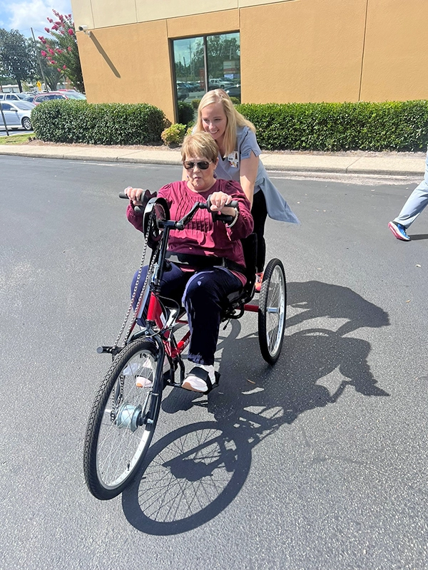 woman in red sitting in a handcycle in a parking lot. A woman in a gray polo is helping by pushing her from behind