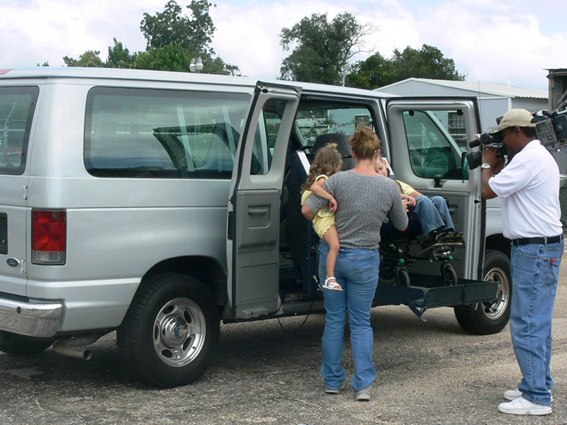 Young Danny and his wheelchair are lifted into his family's van while a camera man, and his mother holding his little sister supervise