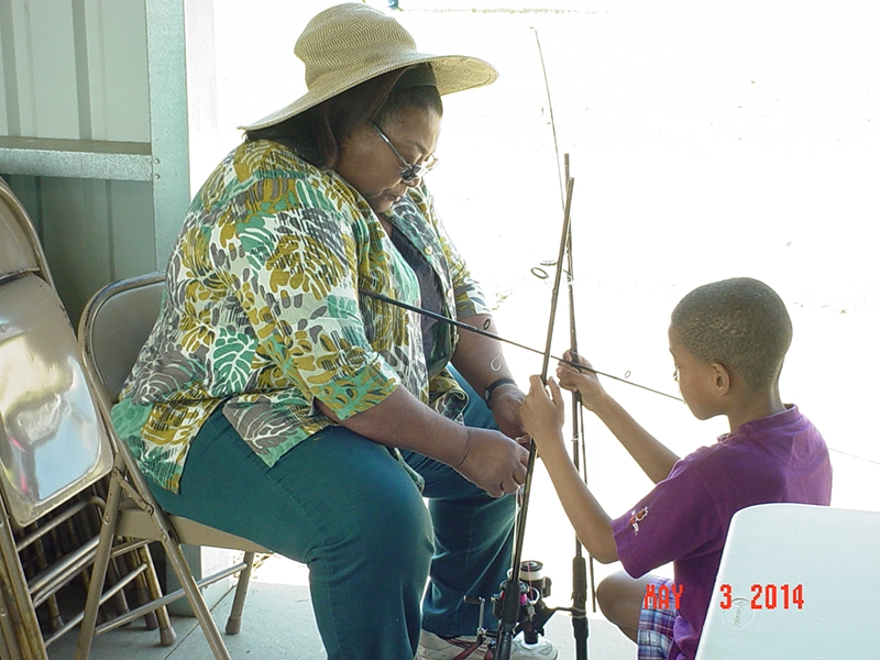 woman in a hat holding and setting up a fishing rod. A young boy in a purple shirt is crouched down facing her while holding up two more fishing rods