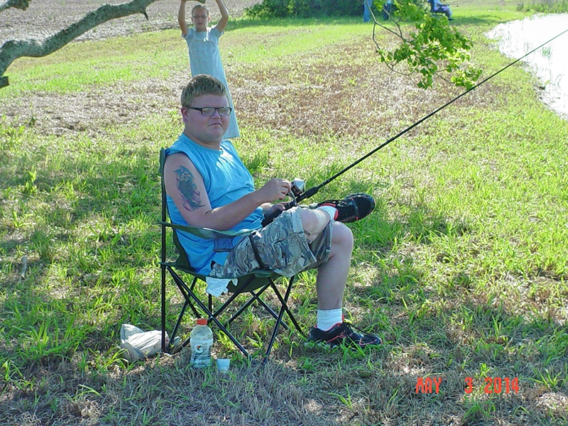 young participant of Penwheels Fishing Rodeo sitting in the shade in a camping chair with a fishing rod pointed toward the lake