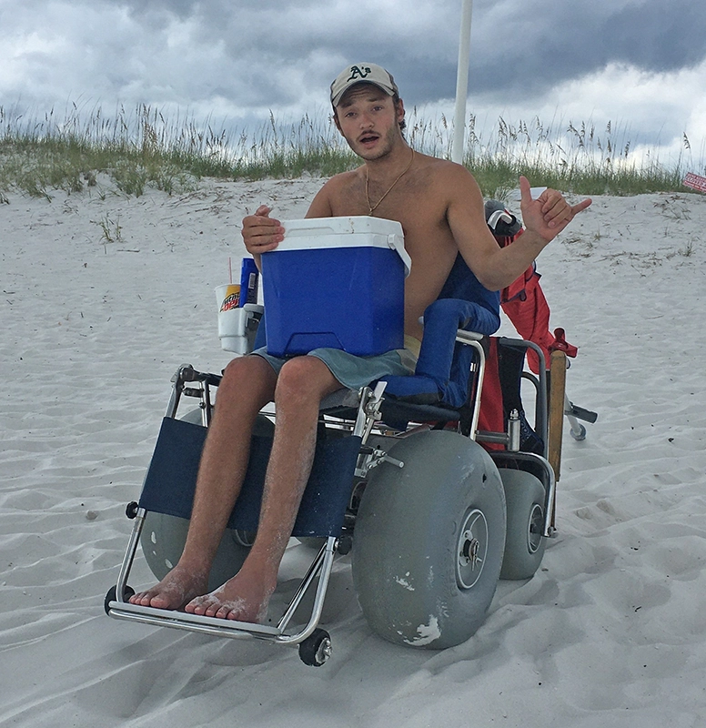 Elis sitting in a wheelchair with thick wheels that can travel easily over sand. Elis is giving a "hang loose" sign and holding a cooler, ready for a day at the beach