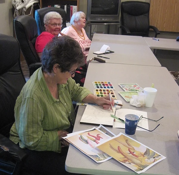 Three senior ladies sitting at gray tables. The lady sitting closest to the camera is painting with watercolors