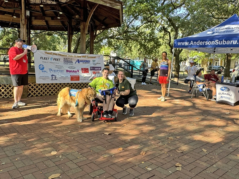 the awards ceremony after a 5k running race events where this competitor competed using a hand cycle in the handcycle division created so people with disabilities can be included to participate. This photo is after the race when the competitor is back in her wheel chair and she is accompanied by her service animal. 